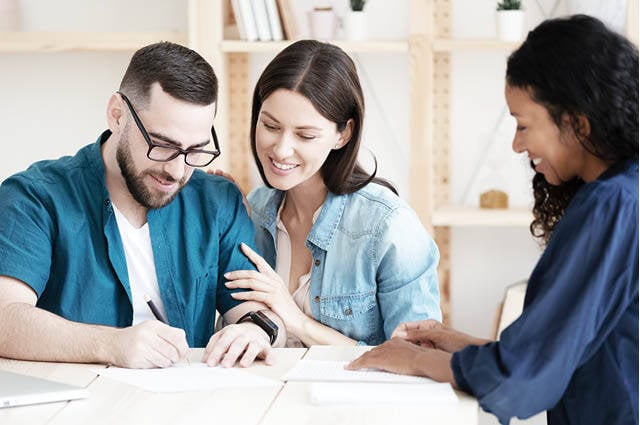 Young couple sits smiling at a table and is signing documents in front of encouraging woman. 