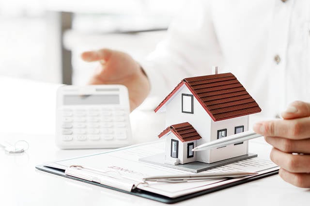 Image featuring small model house and calculator with man's hand in the background.