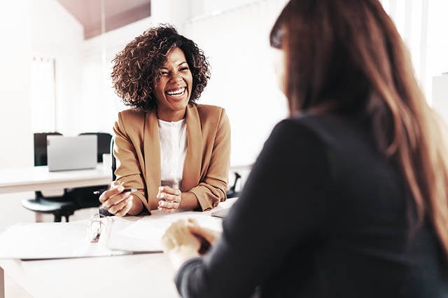 Image with two professional and smiling women sitting across from each other in an office. 