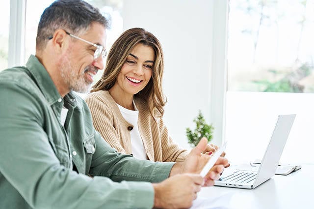 A man and woman sit side by side at a table smiling and looking at paperwork.